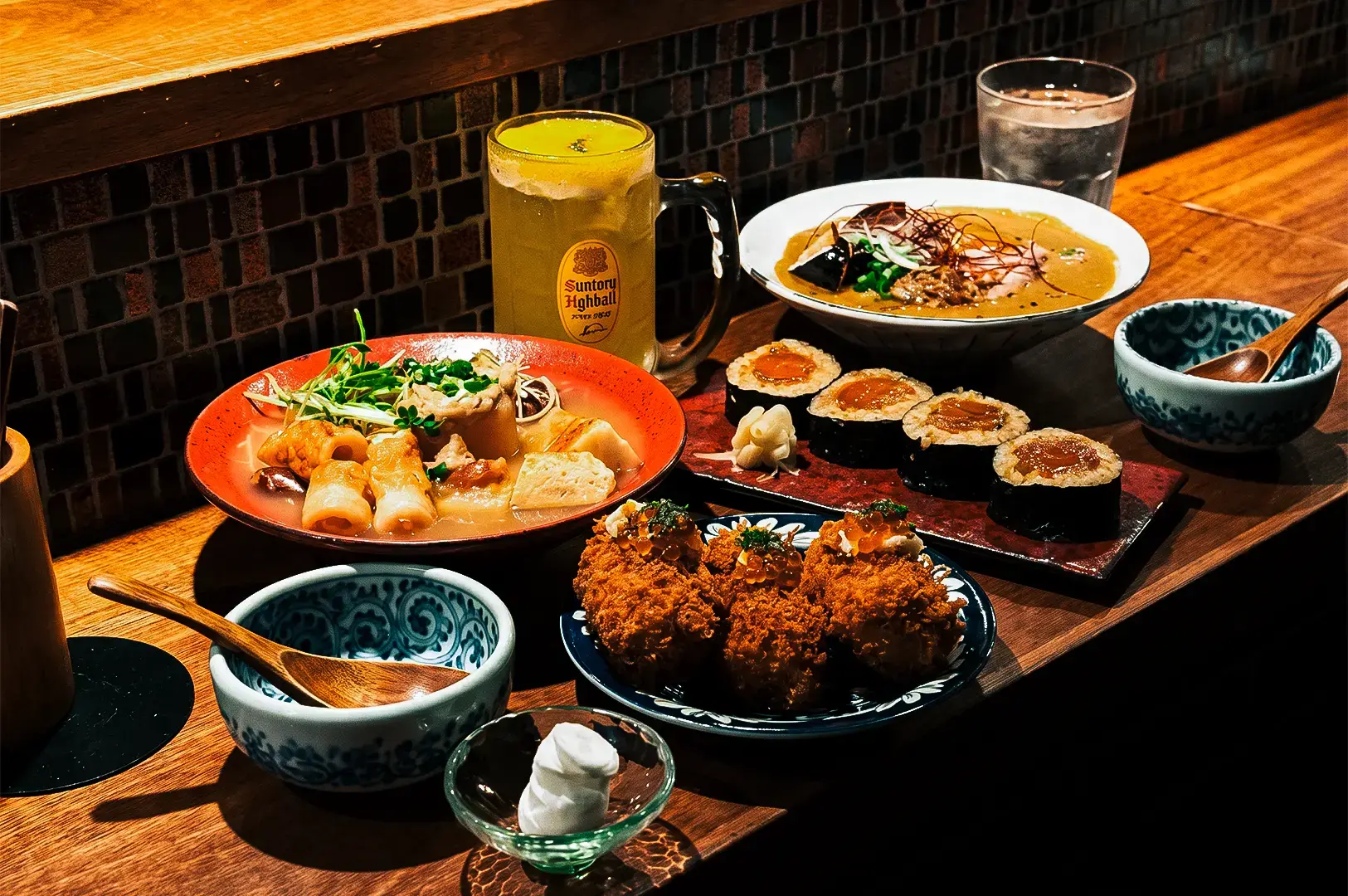 Apollon AM-Bar famous menu dishes Egg-Yolk Maki, Crab Croquettes, and soup dishes arranged on a wooden table together with side bowls and glasses beside the counter.