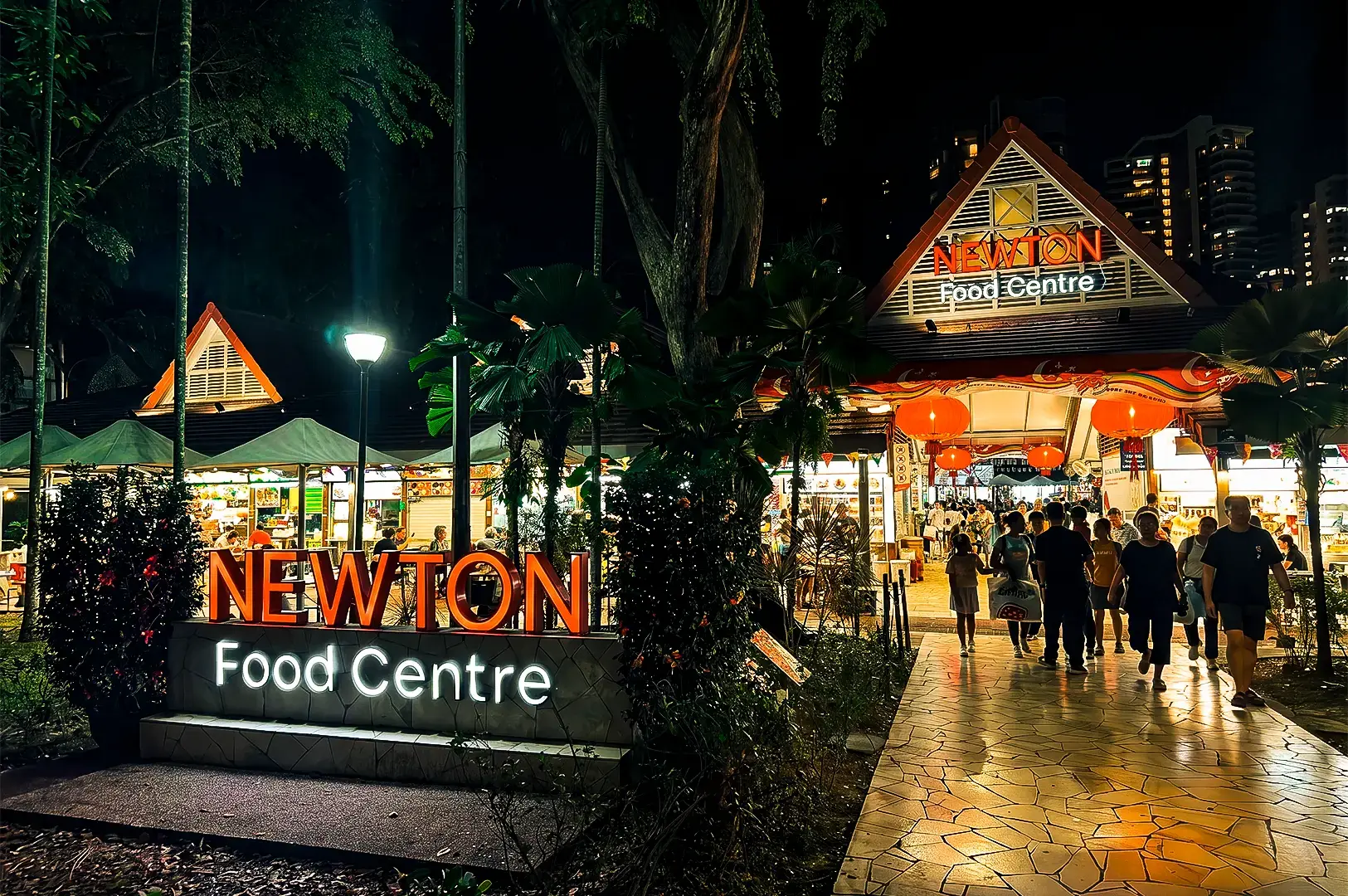 A wide view of Newton Food Centre at night, with its red and white logo in front and above the main entrance.