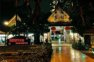 Newton Food Centre at night, with its arched black and red signage at the front and the interior visible behind illuminated by bright white lights