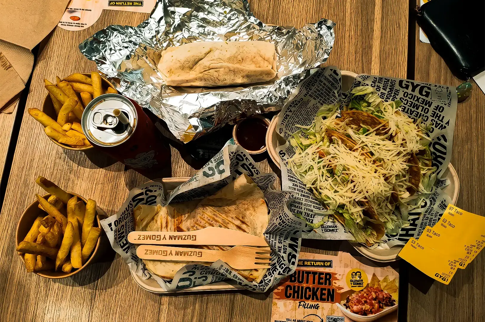 Flat lay wide shot of a variety of Guzman y Gomez menu items including tacos, fries, sauces, and sodas arranged on top of a wooden table.