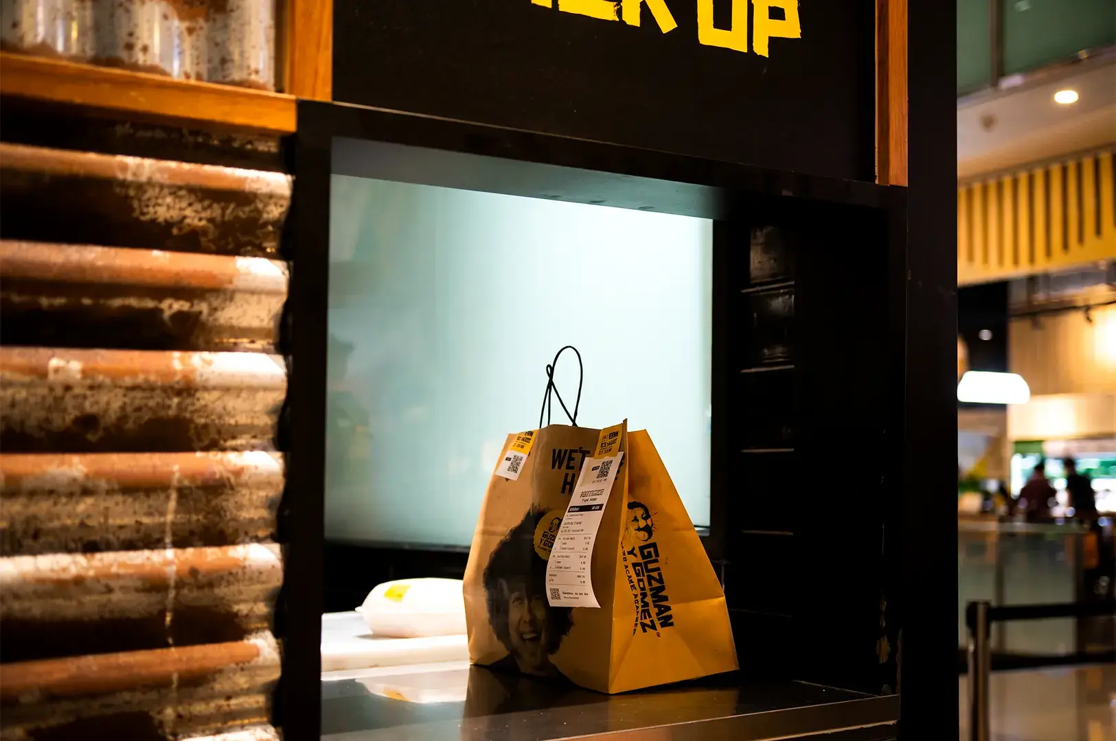 A lone Guzman y Gomez takeout paper bag placed on top of a black counter with wooden wall by the side and the busy area behind.