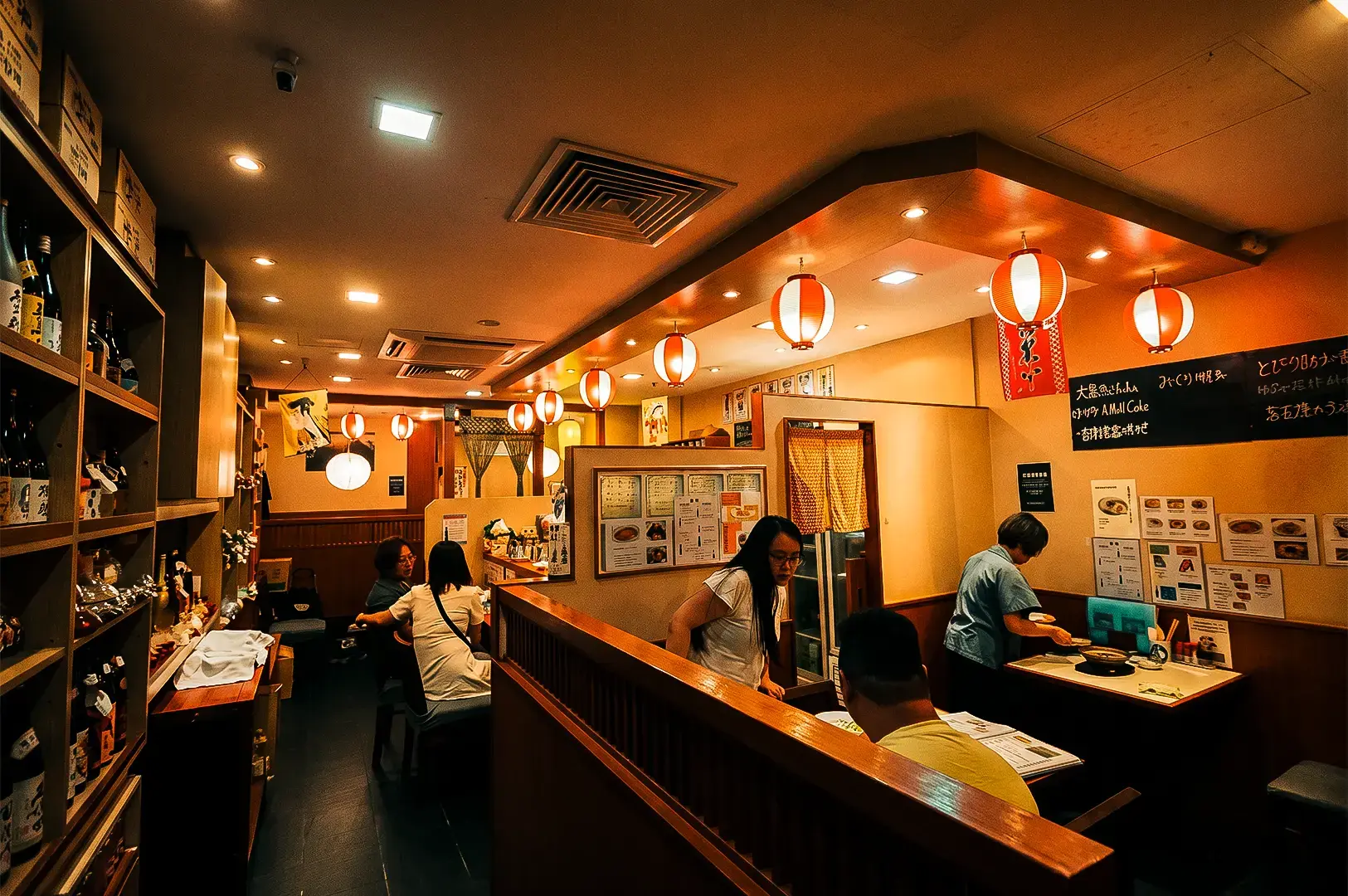 Keria Japanese Restaurant interior with shelves full of bottles on the left and tables and chairs with customers around the interior illuminated with warm yellow lighting from hanging lanterns.