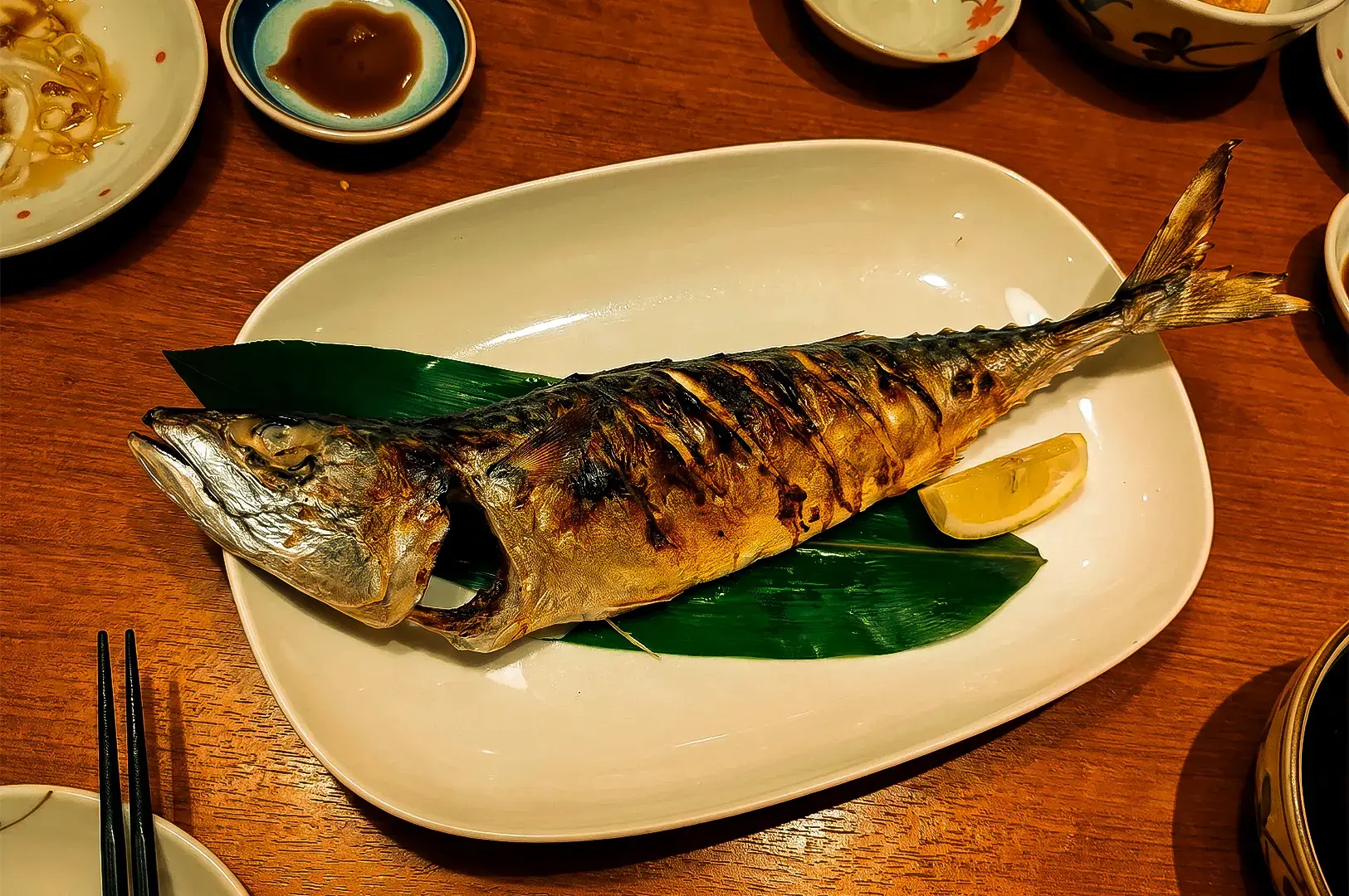 Keria Japanese Restaurant's grilled fish on top of a white plate with a green leaf and lemon slice together with other plates and sauces arranged on top of a wooden table,