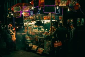 A night market street food stall contrasting its dim evening background with its warm and cozy lights and signages and smoky street foods.