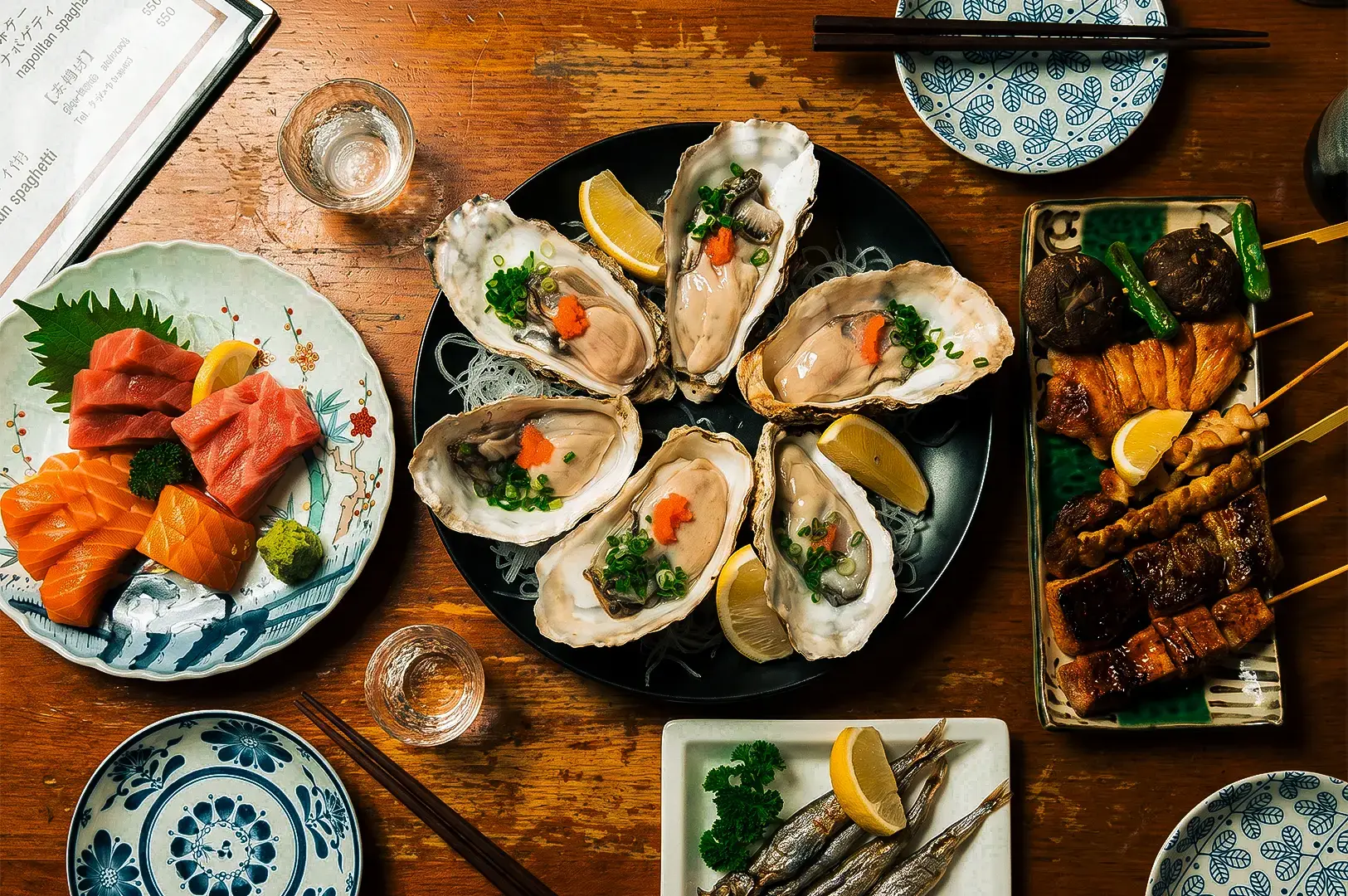 Flat lay shot of Keria Japanese Restaurant menu items with oysters, sashimi, and grilled dishes on platters arranged on top of a wooden table.