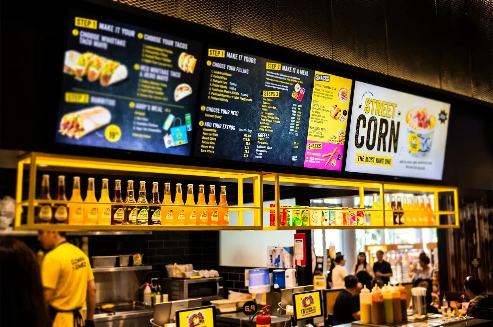 Guzman y Gomez digital menu boards illuminating its store front with white and yellow lights, the workers behind the counter busy preparing orders and various sauces and drinks displayed by the counter.