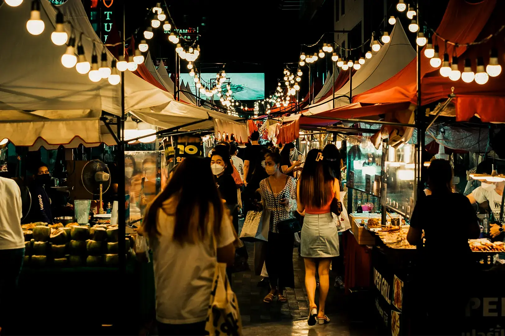 Wide shot of a night market with vendor stalls on either side and customers bustling in the middle street illuminated by warm yellow lights.