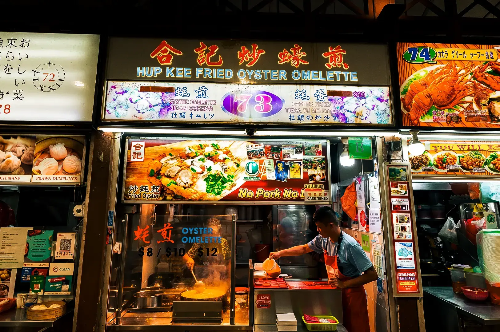 Hup Kee Fried Oyster Omelette stall exterior with brightly lit signages on top and a cook preparing a dish inside.