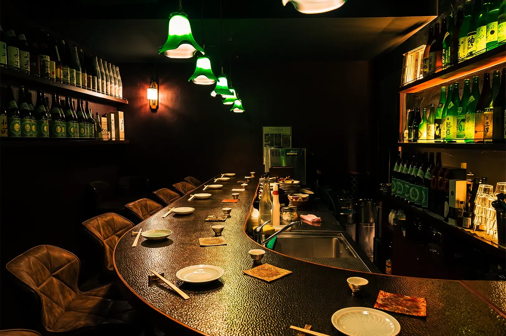Apollon AM-Bar sake bar room interior wide angle shot with sake bottles lined up on a shelf on the right and empty chairs and countertop arranged with utensils and bottles illuminated by warm yellow and green dim lights.