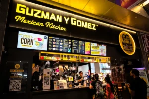 Guzman y Gomez store exterior with its large logo signages and digital menu boards brightly illuminating the area with with white and yellow lights, customers lines up by the counter face a worker taking orders.