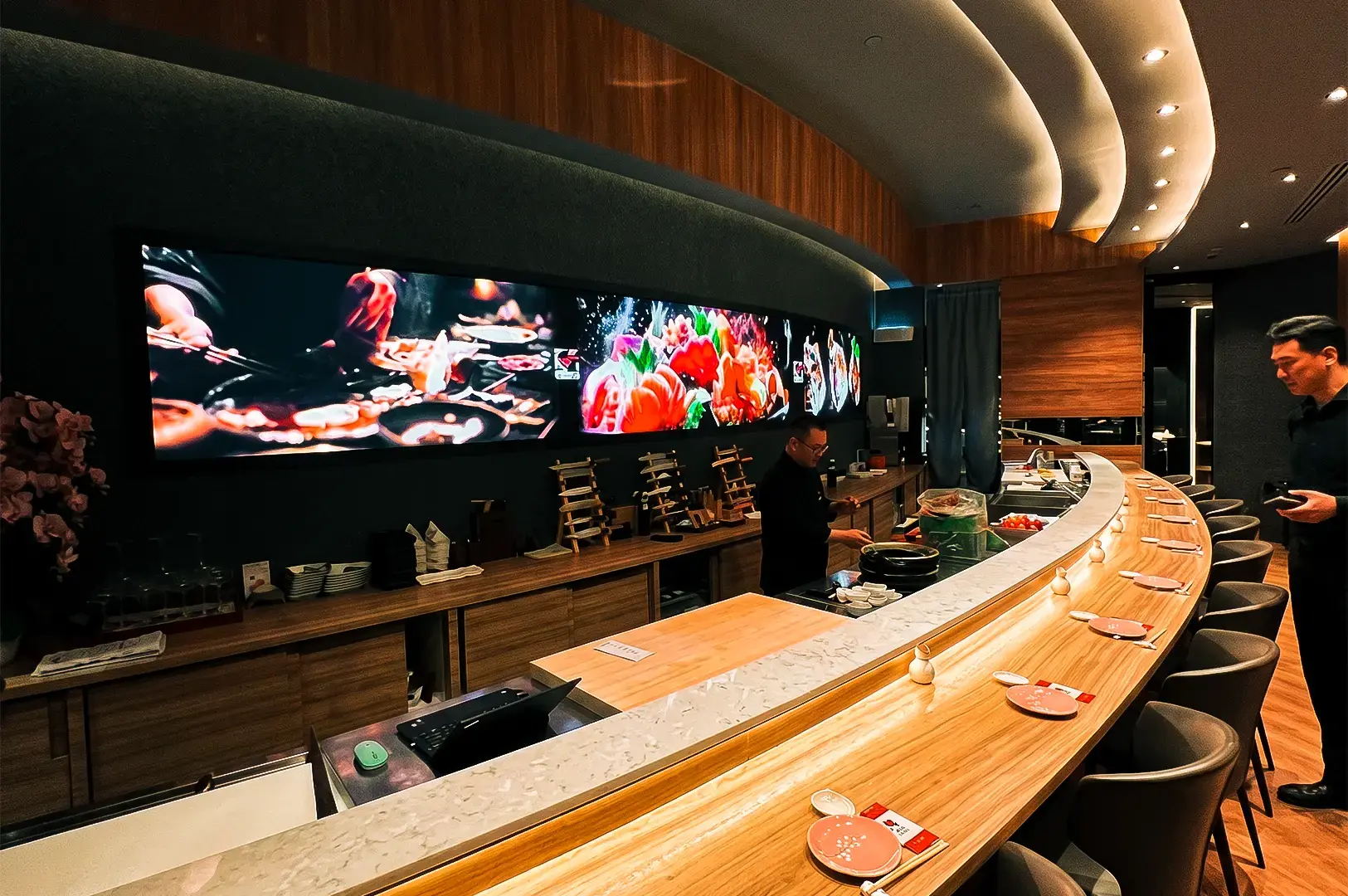 Issho Izakaya interior counter with a line of empty chairs and a staff behind the counter with displays of sushi and other dishes on display on screens.