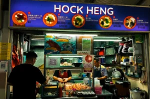 Hock Heng Fish Soup stall exterior with its blue logo signage displaying menu items above the counter with staff preparing dishes inside.